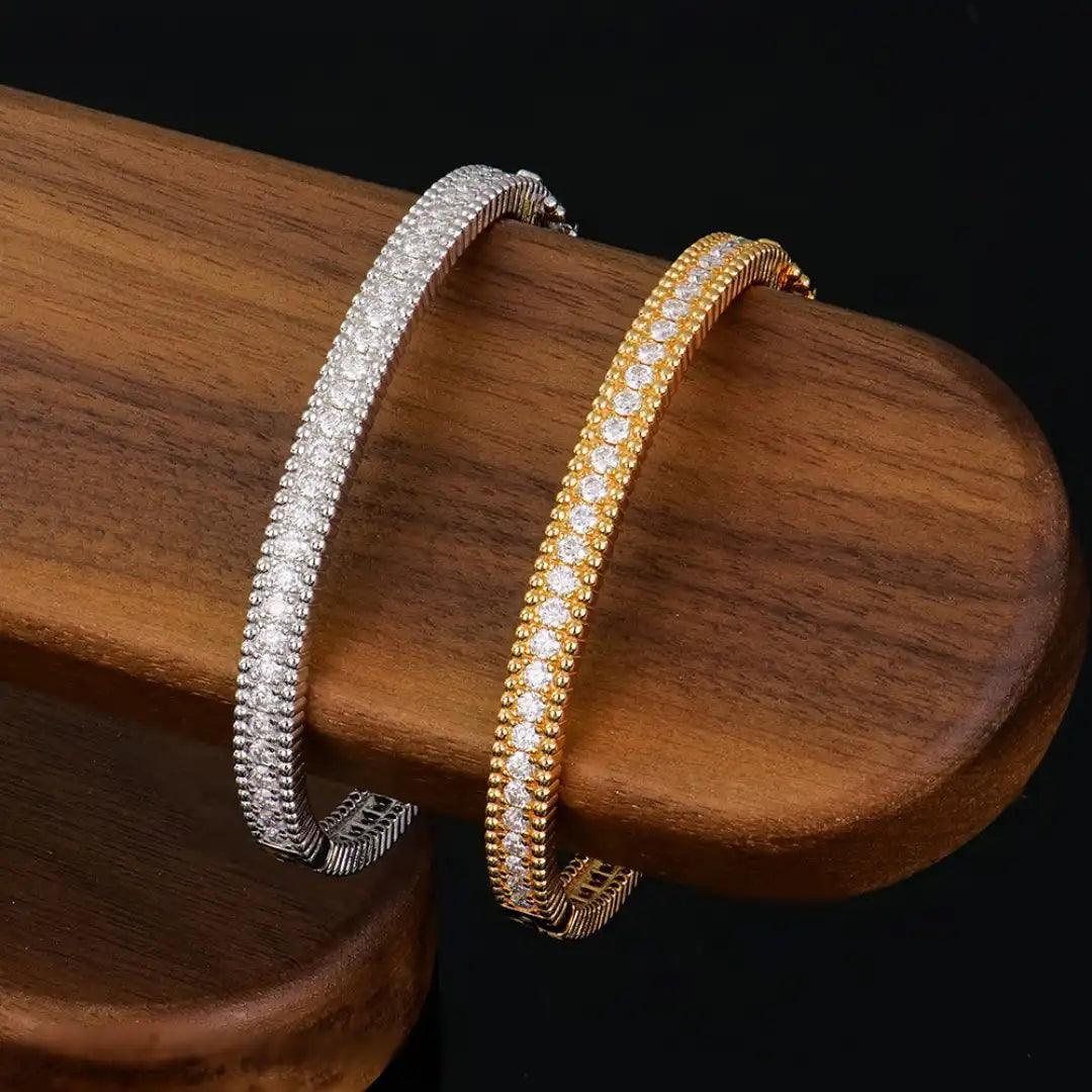 Gold and silver editions of the Mozi Celestial Moissanite Bangle displayed side by side on a wooden platform, showcasing the intricate detailing and sparkling moissanite stones